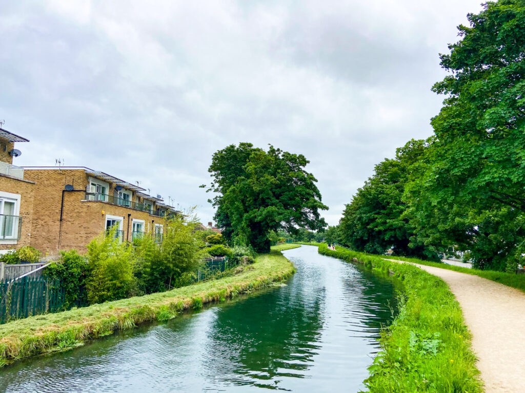 image shows the River Lee canal in Lee Valley, surrounded by some flats on the left and a bike path on the right. Hartfordshire, England in May