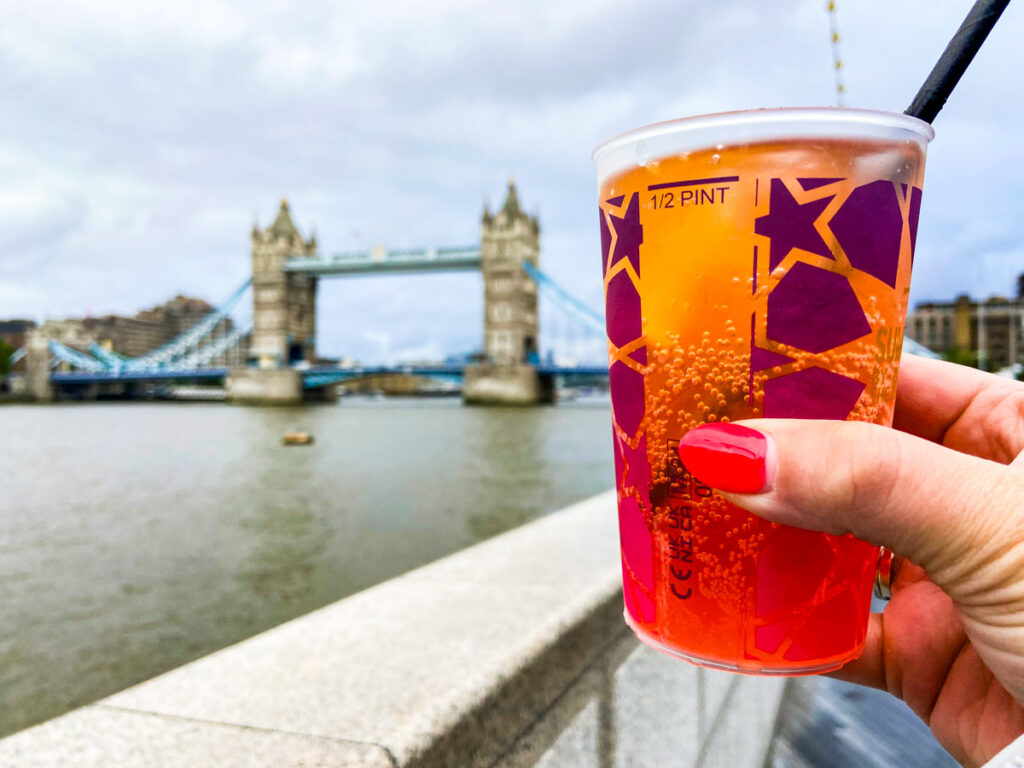 image shows an aperol spritz being held in the foreground against the tower bridge by river thames, taken from summer by the river hidden gem london