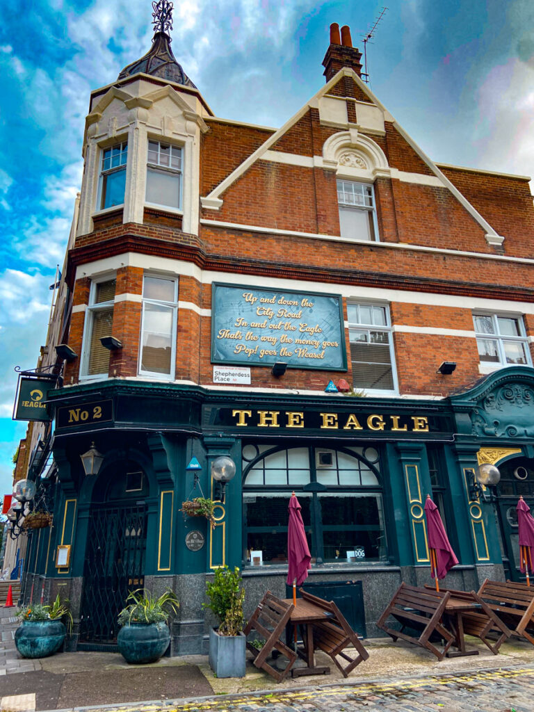 image shows a traditional english pub called The Eagle, with red brick roof and green exterior. Some picnic tables sit empty out front in Hackey, Shoreditch neighborhood of London in May.