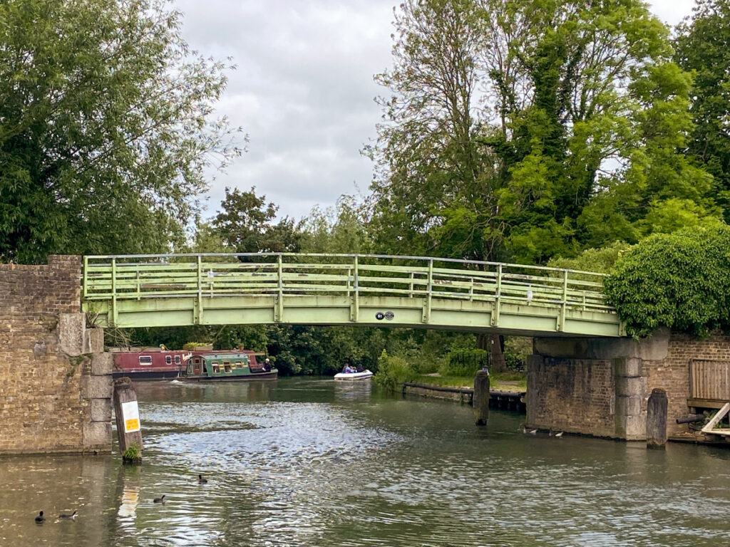 image shows a small green bridge over a river-like canal, with some ducks in the foreground