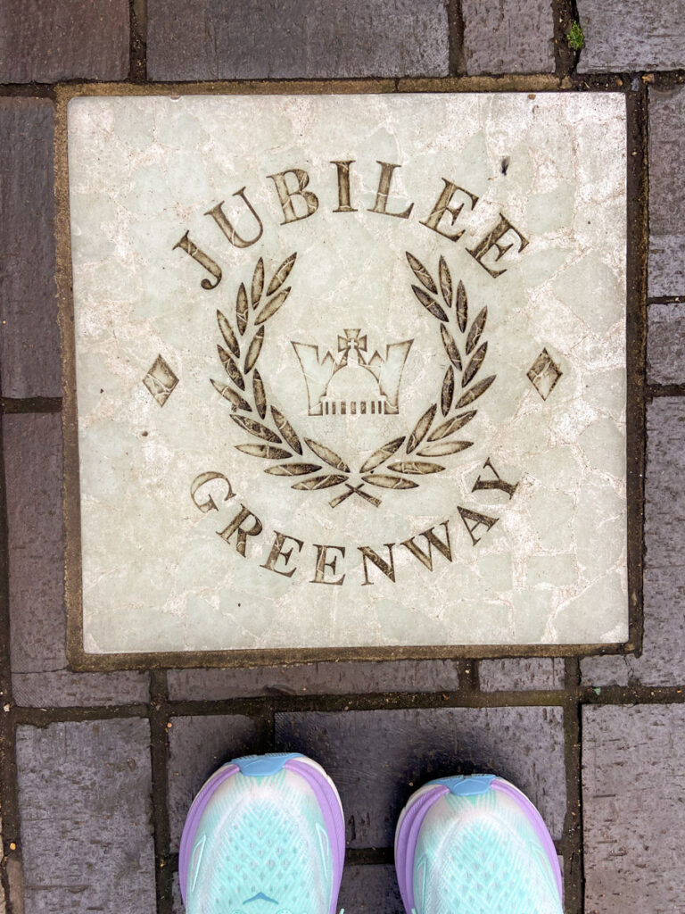 image shows a plaque in the grouund that says Jubilee Greenway with a crown and laurel leaves, and the toes of some running shoes