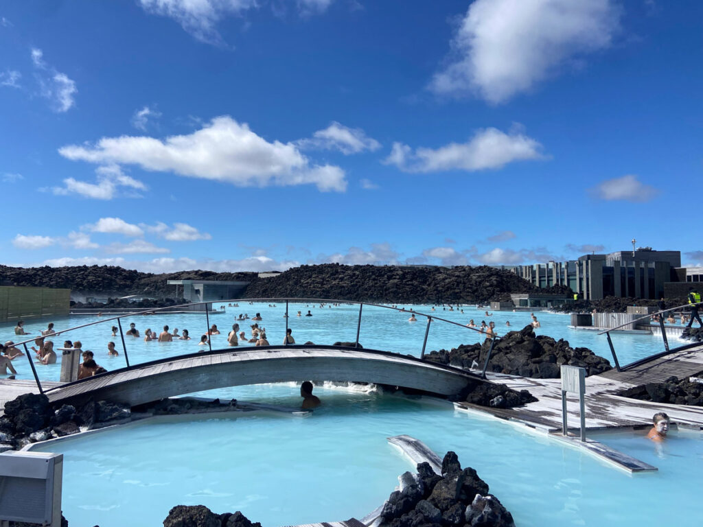 image shows a blue thermal mineral spa with a bridge connecting two sides, with lava walls in the background