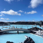 image shows a blue thermal mineral spa with a bridge connecting two sides, with lava walls in the background