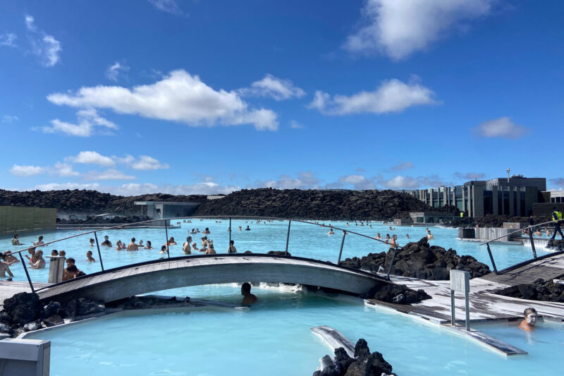 image shows a blue thermal mineral spa with a bridge connecting two sides, with lava walls in the background