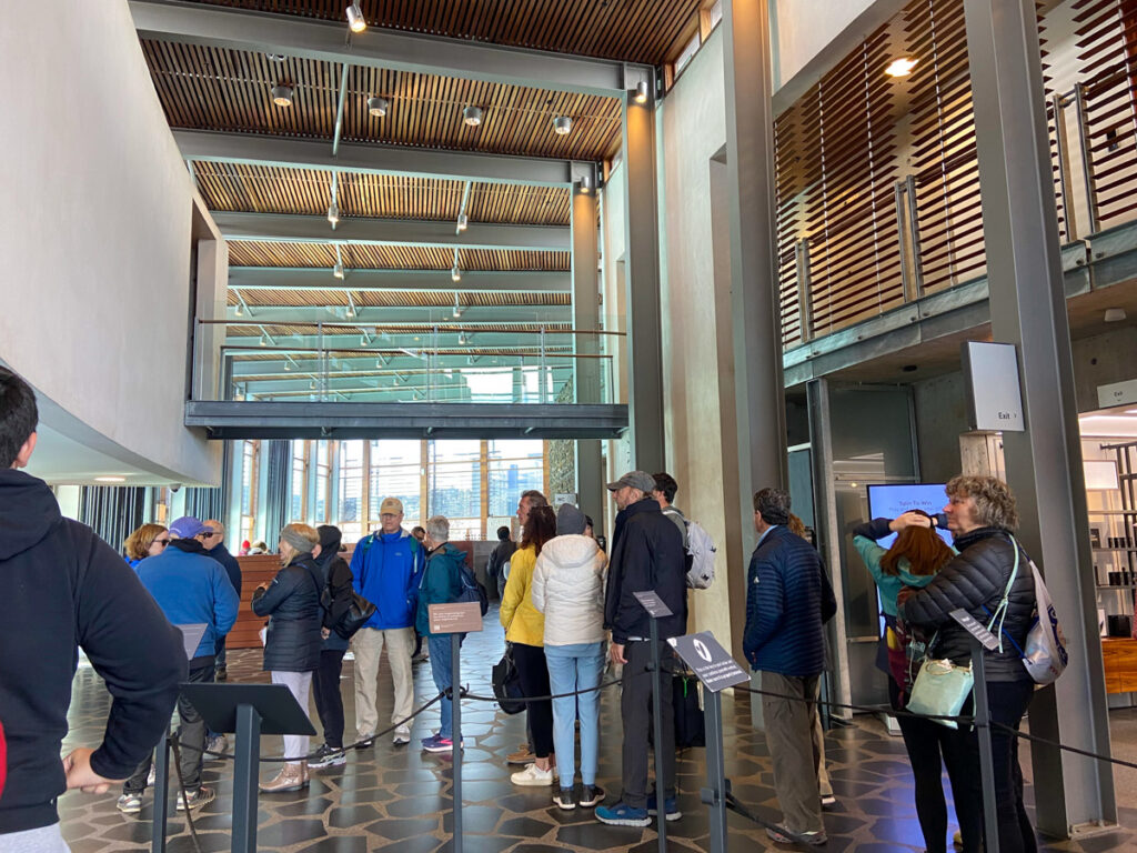 image shows a line of people waiting in a large lobby with a high ceiling