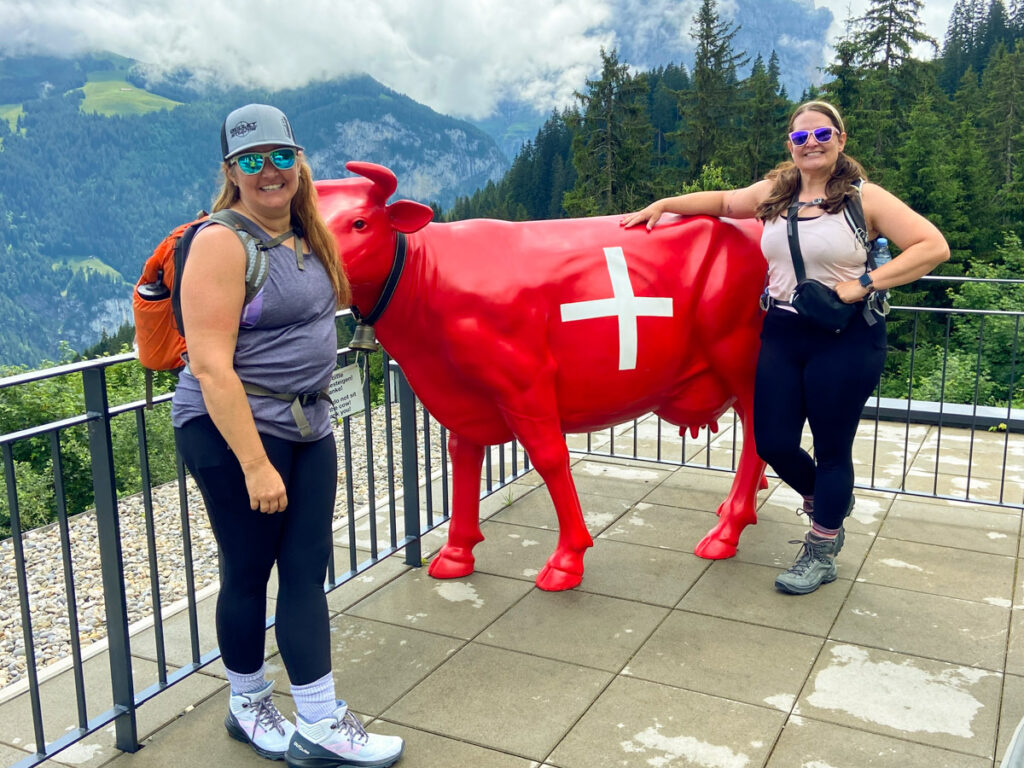 image shows two women in hiking gear standing in front of a painted statue of a cow. the cow is painted to look like the swiss flag. in the background are the jungfrau mountains