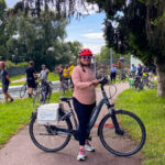 image shows a woman in a pink shirt and red helmet on a bicycle in front of a canal