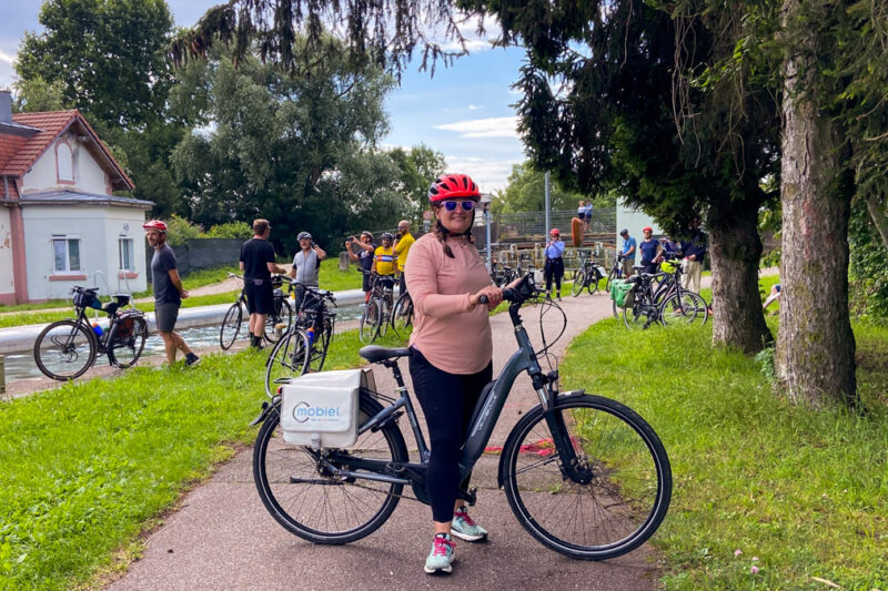 image shows a woman in a pink shirt and red helmet on a bicycle in front of a canal