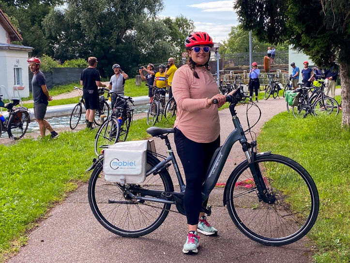 image shows a woman in a pink shirt and red helmet on a bicycle in front of a canal