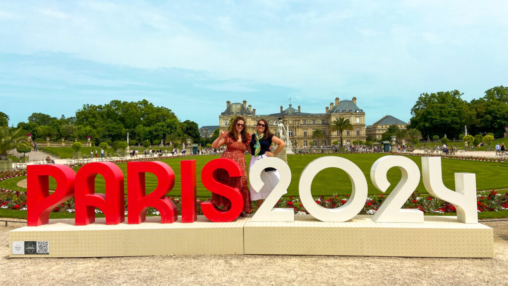 image shows the olympic paris 2024 logo as a large sign with two women standing behind it for a photo