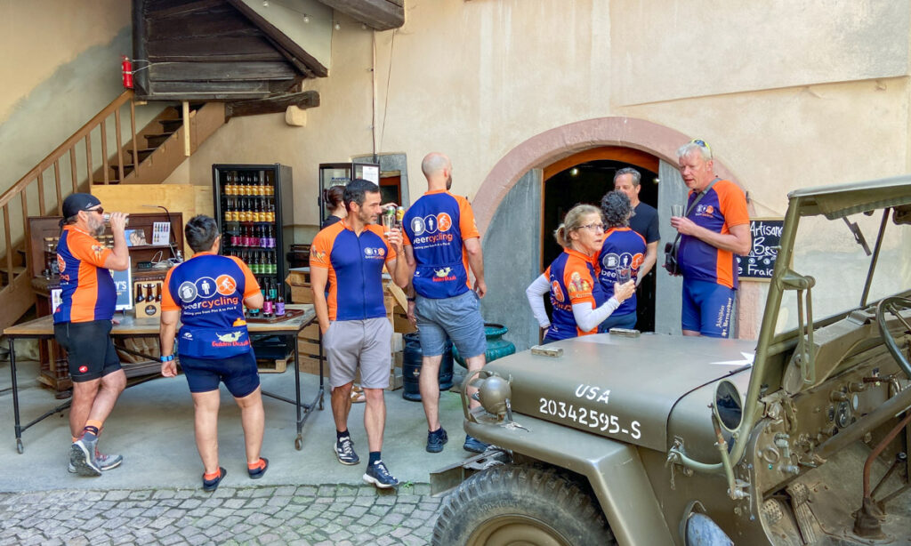 A group of cyclists, all wearing blue cycling jerseys with orange trip mill about a stone courtyard. Many are drinking beer tastings. A WWII jeep is in the front right corner as decoration.