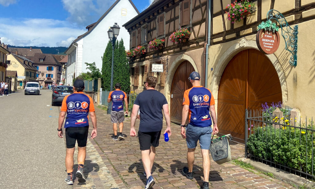 A group of four people walk through the half-timbered buildings of a medieval looking French town. Three of the men are in blue and orange cycling jerseys. 