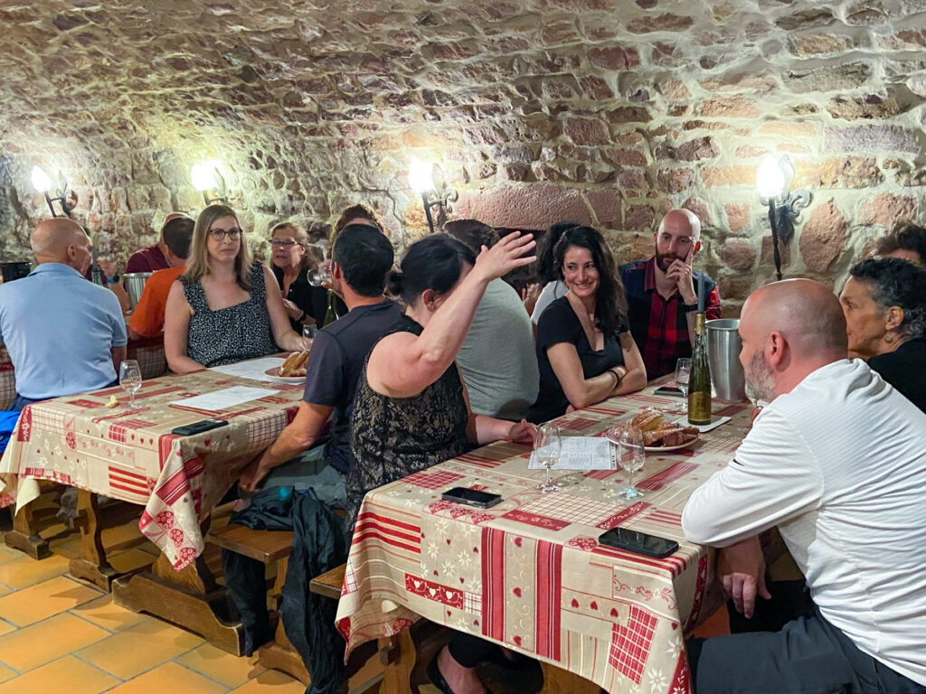 The wine-tasting room at Scherb Winery, nestled in a centuries-old stone cellar, showcasing traditional Alsatian architecture. The stone walls are curved. Three tables of people are in front, with people drinking wine and eating cakes.