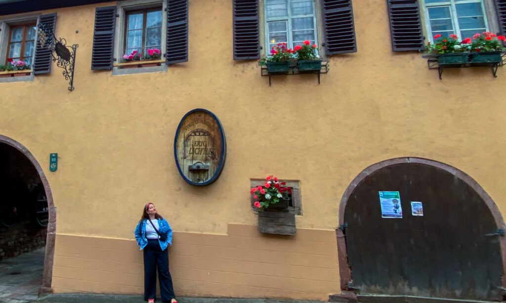 a brunette woman in a jean jacket and black slacks stands in front of a large yellow ochre painted wall. The small windows have bright pink geraniums in the window.