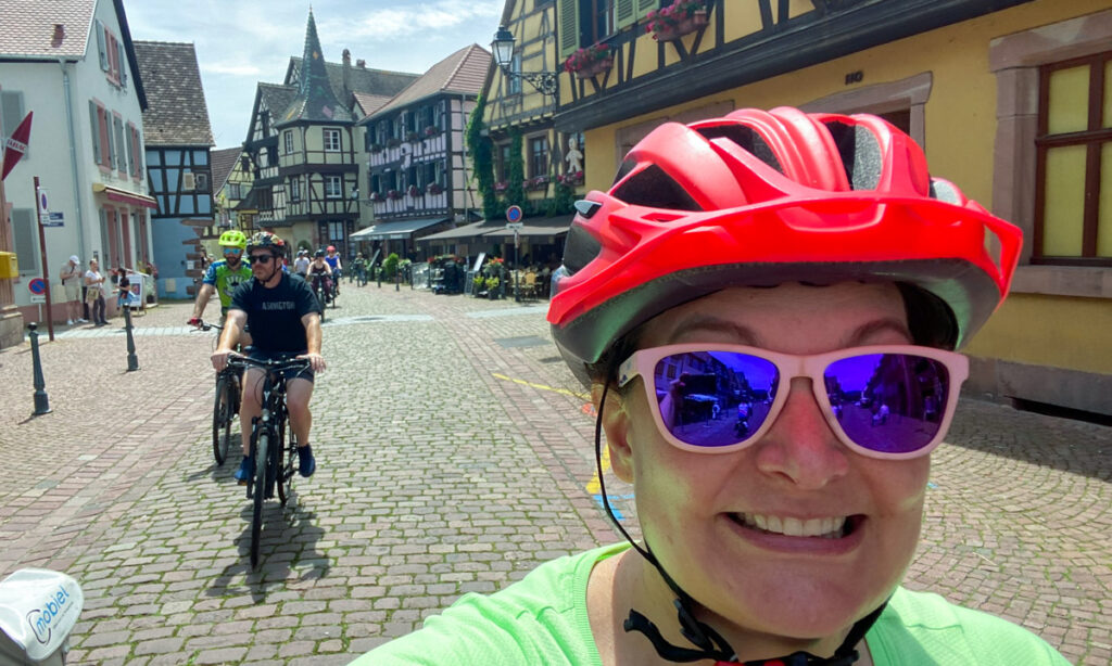 Cyclists riding through a picturesque Alsatian town with half-timbered buildings and cobblestone streets, reflecting the region's medieval charm. The rider in front has a neon orange helmet, pink sunglasses and a brought green shirt.