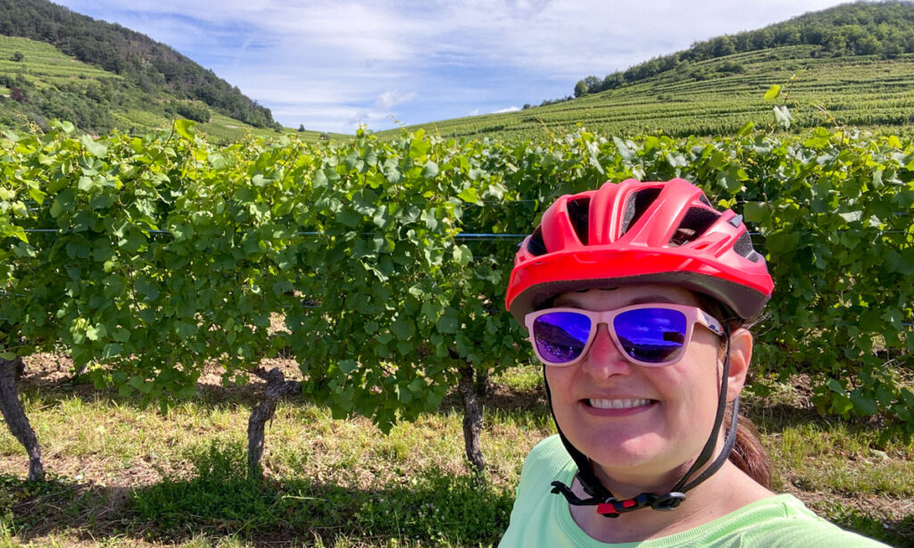 A woman in a bright orange bike helmet, pink sunglasses and bright green shirt stands in front of grape vines. 