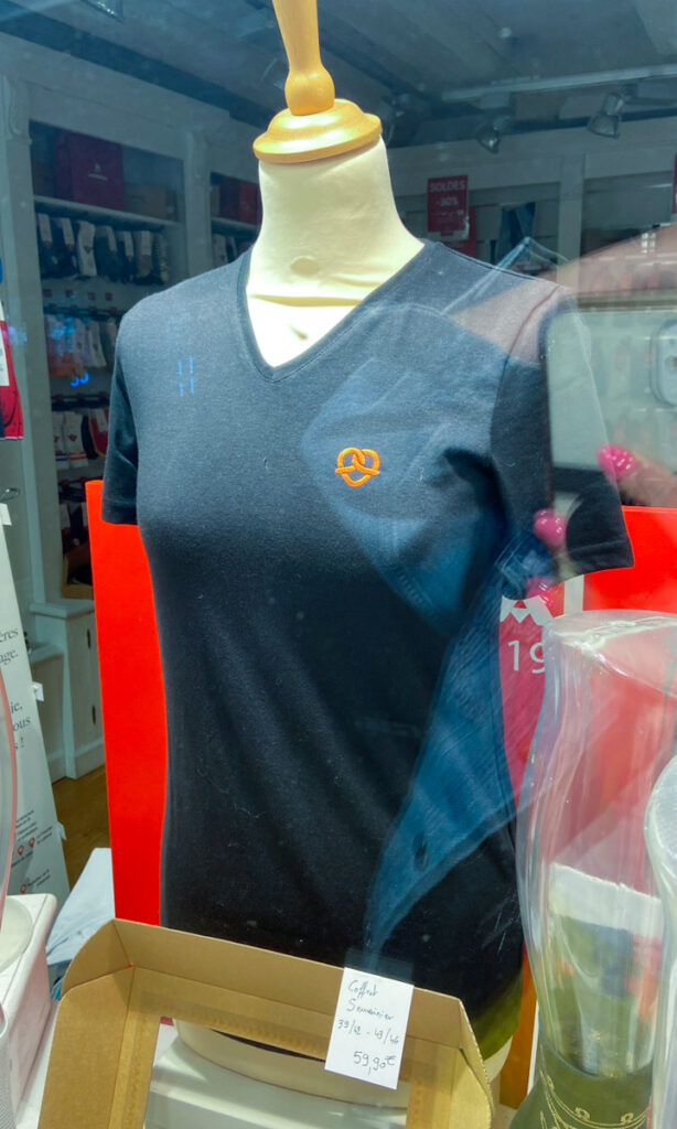 A black t-shirt featuring a large pretzel design, the logo of Alsace. The tshirt is on a mannequin in a shop window.