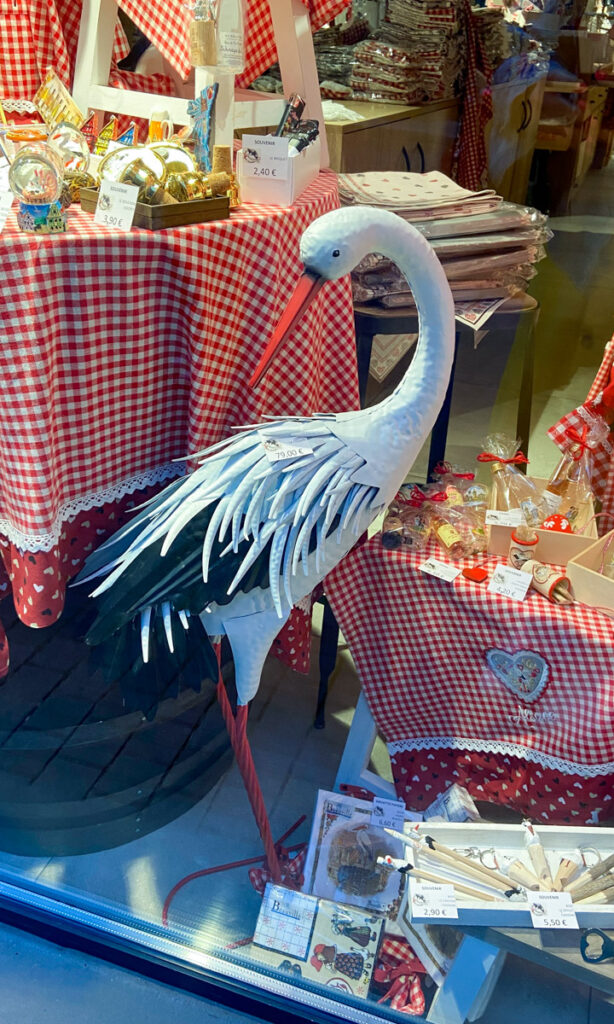 A metal sculpture or statue of white stork is in a store window. ALso in the window are red checked table clothes with snacks and souvenirs piled on top.