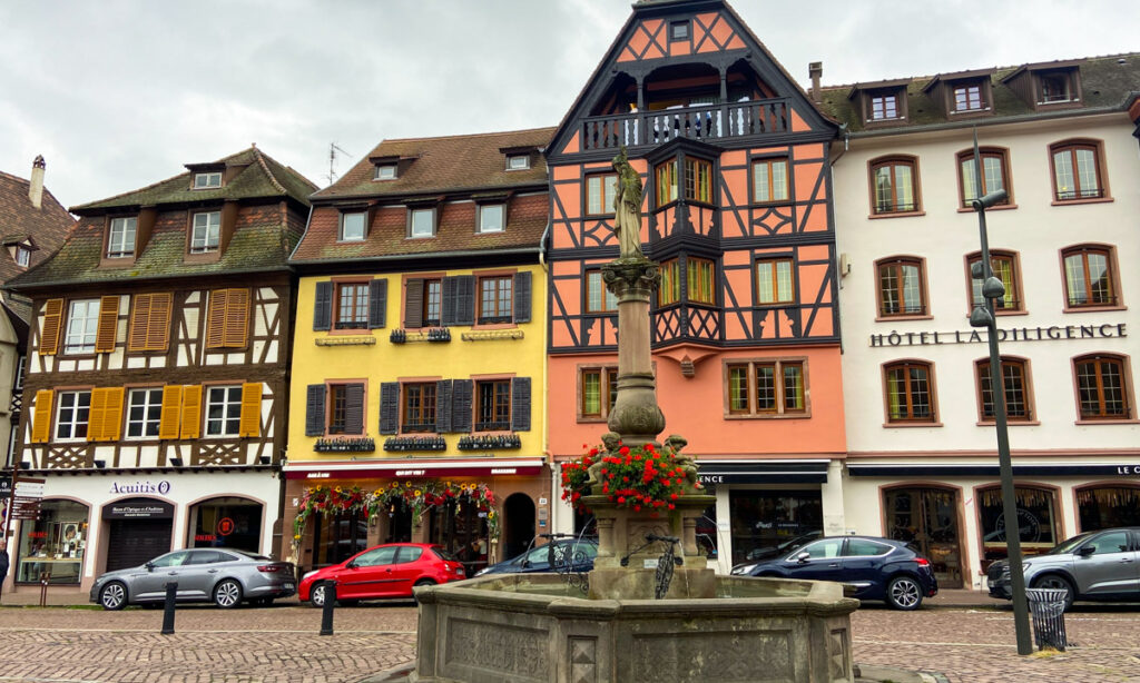 A quaint Alsatian town with half-timbered buildings and brightly painted walls. In front is a fountain.