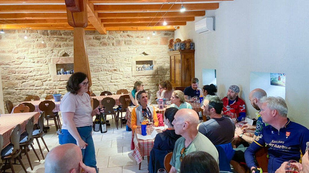 A group of people sit around tables in a tasting room, drinking wine. A woman with short brown hair and a pink tshirt guides the tasting while leaning against a pillar