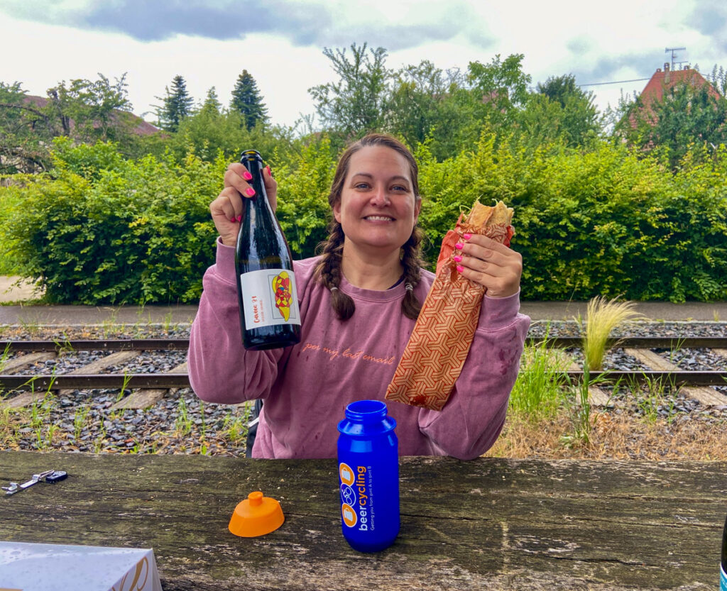 A woman in a pink sweatshirt holds a bottle of wine in one hand and a baguette sandwich in the other. a Blue water bottle is in front of her. She sits at a picnic table outside.