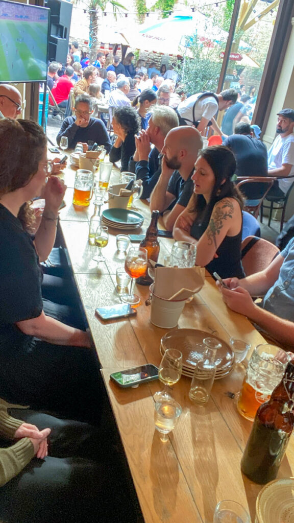 A group of 16 people sit at a very long table, which has numerous beer steins and glasses on it. The photo is taken at a high angle to see everyone at once.