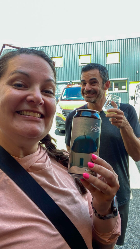 A woman in a pink shirt holds up a can of beer, while a man stands behind her holding up a tasting glass of beer.