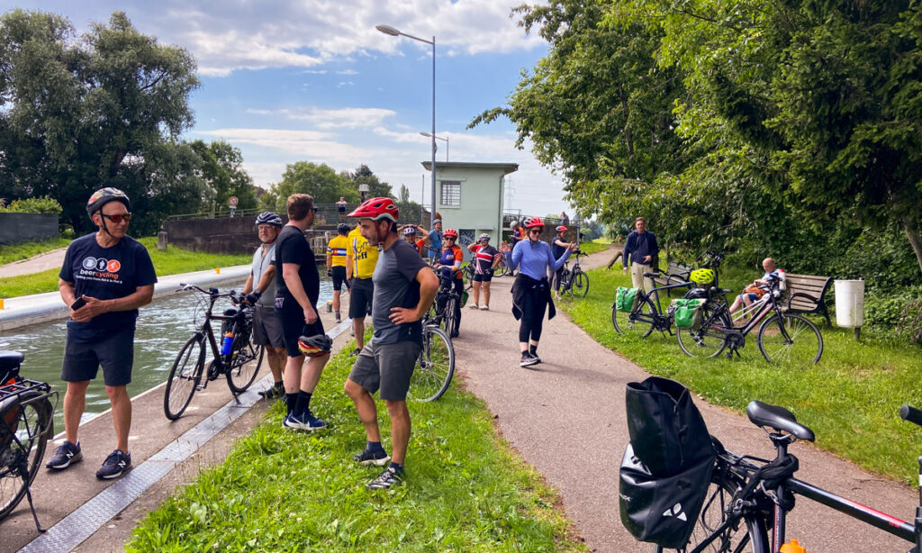 The cycling group stops for a break along the canal towpath, bikes parked beside them, with an easy, paved path visible.