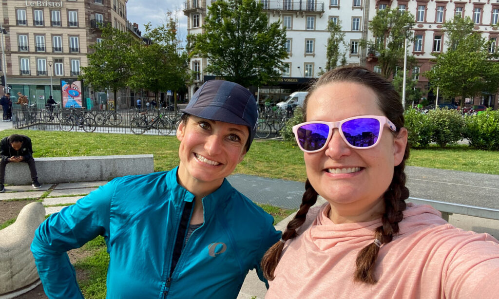 two women smile at the camera. One on the left has a navy cycling hat on and a blue jacket. The one on the right is taking the selfie and has pink sunglasses, and a pink shirt on. 