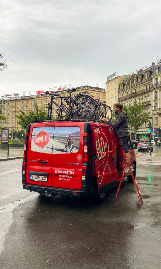 Image shows a red sprinter van that says Duvelo on it, with several bikes on the roof. A man on a small stepstool arranges the bikes on the roof.