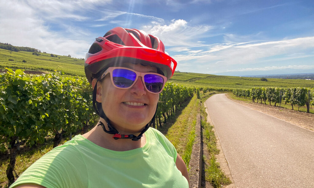 a woman in a bright orange cycling helmet, pink sunglasses and green shirt stands in front of a country road lined with grape vines. 