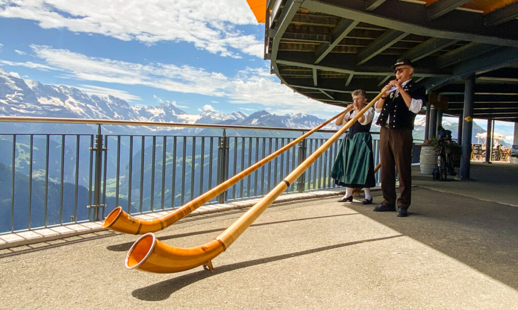 image shows two people in traditional Swiss dress blowing alphorns at the top of Schynige Platte, with mountains in the background