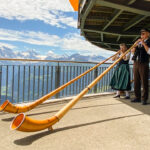 image shows two people in traditional Swiss dress blowing alphorns at the top of Schynige Platte, with mountains in the background