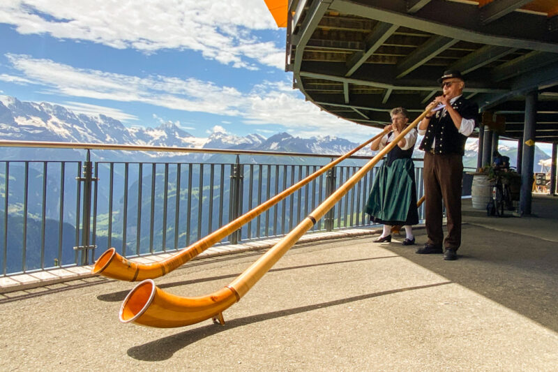 image shows two people in traditional Swiss dress blowing alphorns at the top of Schynige Platte, with mountains in the background