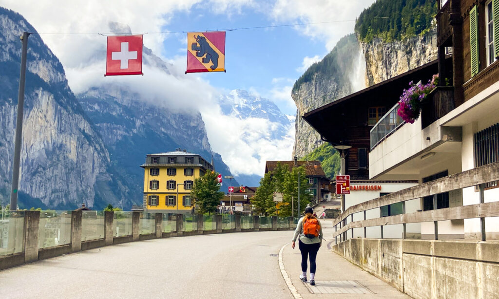 image shows a scenic vista of mountains on the left, a yellow hotel, and swiss buildings on the right. in the background is a waterfall. in the foreground, a woman hiking with an orange backpack walks down the road.