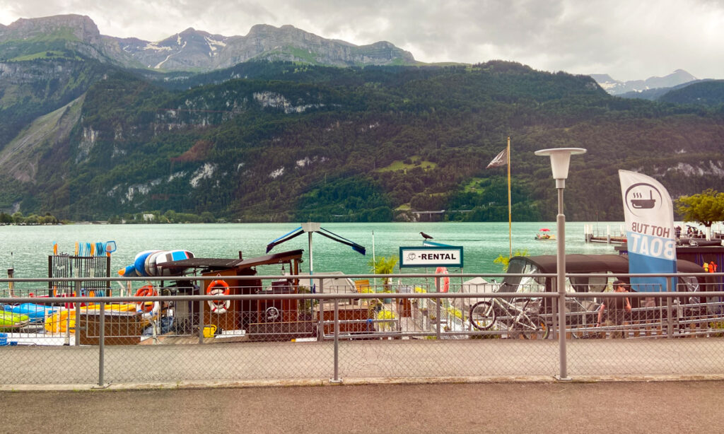 image shows mountains in the background, with a greenish teal lake, and then a mishmash of items along the shore, including boats, bikes, and a sign that says Hot Tub Boat